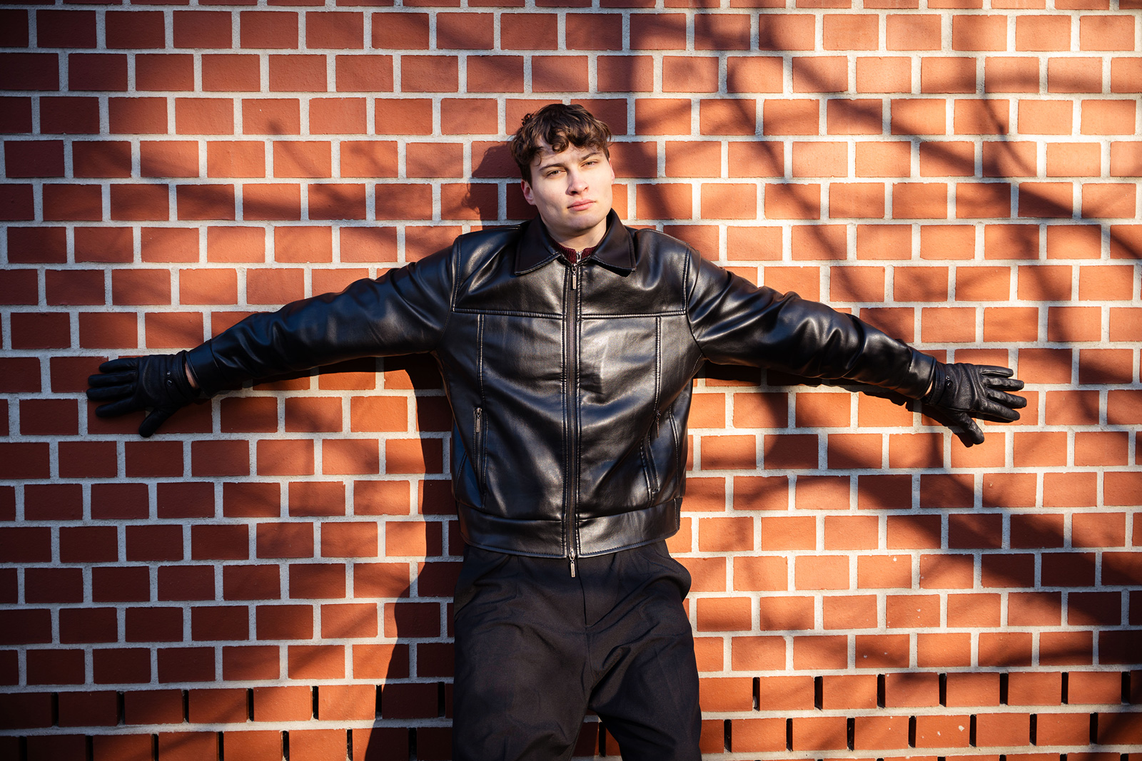 Tobi lehnt an einer roten Backsteinwand, hinter ihm die Schatten eines Baumes an der Wand